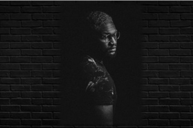 A black and white photo of a man in front of a brick wall.