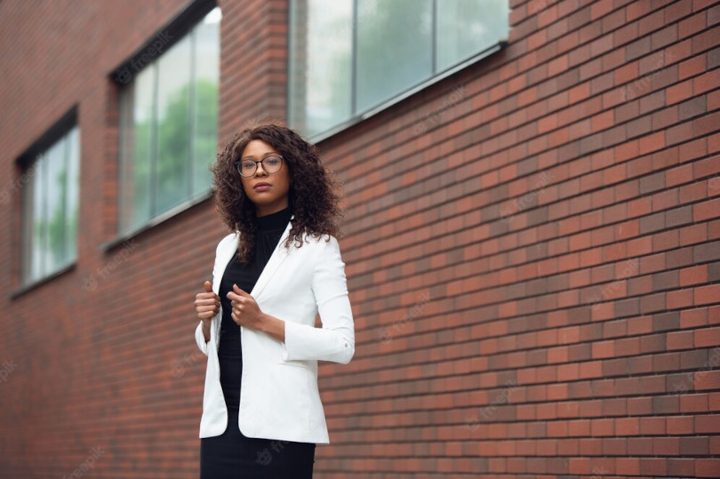 A woman in white jacket standing next to brick wall.